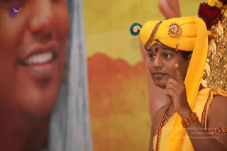 A man in a yellow turban with a teal-and-gold ornament and a red bindi stands in front of a large printed portrait of a smiling face on the left. He wears a yellow shawl over an ochre-and-gold robe and multiple red rudraksha malas and red beaded bracelets. He raises his index finger in a teaching gesture while looking to the side. Behind him is a gilded ornate seat decorated with red roses. The background is a warm yellow and orange backdrop.