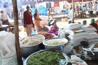 Outdoor relief kitchen under a striped canvas tent with blue tarpaulins, where volunteers prepare food for an annadaan meal. In the foreground, large steel pans hold chopped green leafy vegetables, white grated coconut or rice, and red-orange chilli or tomato preparations, surrounded by sacks of rice and flour. Several women wear white head caps and aprons; one woman in an orange-yellow sari stands near the sacks, and another in a pink sari sits chopping vegetables. A standing man in a red shirt and white dhoti stands beside a steaming vessel. Plastic chairs and tables are visible in the background among other tents and people in white.