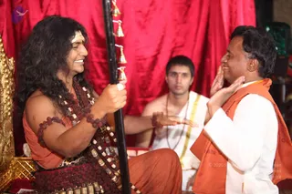 A seated man with long, dark, swept-back hair and a white tilak on his forehead wears orange clothing and multiple rudraksha malas with bead ornaments, holding a black staff hung with small brass bells. He smiles warmly as he faces a shorter man in a white kurta and orange vest who has his palms raised near his cheeks in a gesture of reverence. Behind them a young man in white dhoti with a gold border sits in front of a red draped curtain, and a golden throne-like seat is visible at left. a person: the long swept-back hair, orange clothing and layered rudraksha malas are the distinguishing cues for the seated central figure.