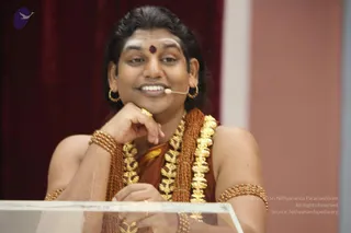 A smiling man with long dark hair sits behind a clear acrylic lectern in front of a deep red curtain. He wears a headset microphone, a white vertical mark and red bindi on his forehead, gold hoop earrings, a brown-orange saffron-toned garment, layered rudraksha malas and a gold ceremonial necklace, and beaded bangles on his wrists and upper arm. His right hand rests under his chin in a relaxed discourse pose.