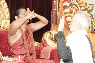 A photograph of two people in a temple-like setting with a golden throne and red zebra-striped cushions. On the left, a man with dark hair, a red bindi and a rust-red shawl over bare shoulders wears a rudraksha-style beaded necklace and sits on the red throne, his hands raised before his face in a mudra gesture. On the right, an older white-haired, white-bearded man in a white shawl and dark grey jacket raises one palm toward him in greeting. Behind them stand a golden deity murti and a backdrop of marigold and white flower garlands, with gold-embossed ornaments and a dark red curtain.