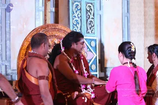 A photograph inside an ornate temple-style hall with a gilded carved throne and blue and white tiled panels. A seated man with long dark hair, wearing a maroon-red robe with an orange edge, a rudraksha-style neck mala and a garland of pink and white flowers, extends his hand while a woman in a pink top with a long braid and white flower in her hair kneels before him, apparently performing padapuja. Another man in a maroon shawl and orange-bordered robe with a beaded armband sits at left, and a woman in a white sari with a red-bordered shawl is at right. The gesture and garland suggest a ritual honouring of the seated figure, and the seated figure's appearance is a probable match to a person, with long swept hair, a garland, and maroon ritual robes.