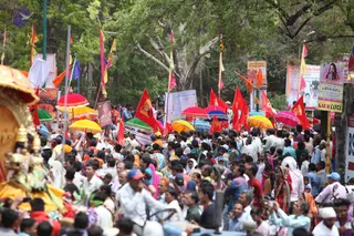 A dense street procession moves along a tree-lined road, with a large crowd of men and women in white, pastel and bright clothing pressing forward. Red and orange flags on tall poles, and many colourful parasols in yellow, red, blue and green, rise above the people. On the left, the gilded edge of a decorated palanquin or ratha is visible, with a painted deity figure. Roadside hoardings and banners show portraits and printed text. Many people are on foot, and a man on a motorcycle is in the foreground.