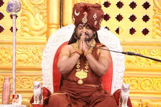 A man seated on an ornate white and red throne with joined palms raised in namaste, eyes closed, in front of a gold-carved backdrop with lattice cutouts. He wears a rust-red turban with a crescent and ornamental brooch, a rust-red robe, heavy gold necklaces, gold bangles on the upper arms and a rudraksha-style bead mala. A microphone stands at his side, a silver staff with a finial is at left, and two silver lion figures flank the throne base. A small brass vessel sits on the left, and a small framed photo appears at lower left. The scene suggests a seated discourse setting.