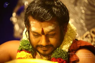 Close-up photograph of a man looking down toward a yellow object in the foreground, with his head bowed in concentration. He has swept-back dark hair, a short beard and white sacred ash marks on his forehead with a red-brown tilak at the centre. He wears a thick garland of white jasmine, green leaves and red roses, and a gold chain, with a rust-orange garment on his shoulder. Behind him, a soft-focus silver and gold decorative backdrop glows. The setting suggests a ritual or initiation session.