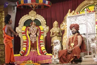 A satsang stage at a temple-style hall with a deity murti dressed in red and gold, adorned with long garlands of yellow, red and white flowers and layered gold-and-white jewellery, seated on a pink-bordered cloth. To its right, a seated man in an orange-red turban and orange robe sits on a leopard-skin cushioned seat before a microphone stand, hands folded, with a gold necklace and rudraksha-style beads. A young man in orange dhoti and bare upper body stands at left, reaching toward the deity with a ritual offering. A golden female statue with folded hands, silver lion figures, a golden horse-head motif and a striped umbrella canopy frame the scene, with fruit set on a tray in front.