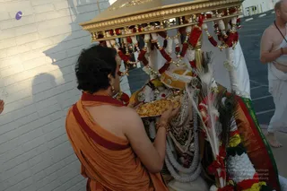 A devotee in an orange saffron garment with a maroon sash stands with his back to the camera, holding a silver plate of golden sweets or offerings up to a richly adorned deity murti seated beneath a golden canopy (palanquin/ratha) hung with red and white flower garlands and bells. The murti wears jewelled silver ornaments and a gold-and-green brocade cloth, with white and peacock-feather fans beside it. Another man in white ritual dhoti and shawl stands at the right edge beside a white brick wall.