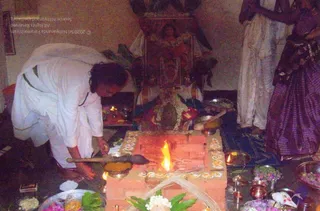 A homa is being performed inside a home shrine room. A man in white dhoti and white shirt kneels over a small brick fire pit, pouring or stirring offerings with a long wooden ladle, while a bright flame rises from the pit. The pit is decorated with white and yellow rangoli-style dots and surrounded by brass plates and bowls holding flowers, leaves, fruit and turmeric. Behind him a deity murti garlanded with white flowers stands on an altar. A woman in a purple striped sari stands at the right edge, and two small oil lamps burn nearby.