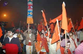 Night procession at the Kumbha Mela with a crowd of devotees carrying saffron-orange flags on tall bamboo poles. A tall painted pole with blue, red and yellow motifs and gold-yellow base rises at left-centre. Many people wear white or saffron clothing, some with white forehead markings and face paint. A man in a tan jacket and white dhoti holds a flag pole in the centre, and a woman in an orange sari with a red shawl holds a flag nearby. Hazy orange street lights and a camera operator at left frame the scene.