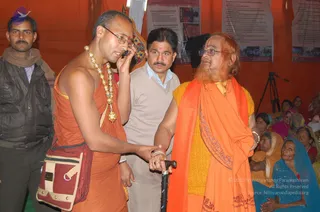 Indoor camp tent with orange walls at a Kumbh Mela event. A bearded elder in an orange shawl over a yellow sweater, wearing glasses and a red tilak, leans on a black cane while a man in a rust-orange sannyasi garment with a large gold bead necklace and a striped cloth bag holds his arm and speaks on a mobile phone. A man in a grey sweater and a man in a dark jacket stand nearby, and women in saris sit behind on the right. Printed posters hang on the orange backdrop. A tripod-mounted camera stands at right. Visible people are roughly seven, so the count range is 6-20.