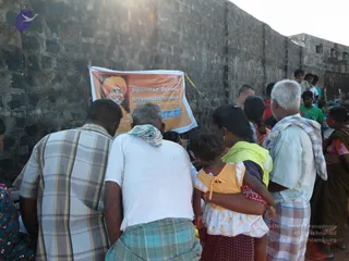 Outdoor scene at a village wall of rough grey stone blocks. A crowd of villagers faces a hanging orange banner showing a smiling man in an orange turban and saffron clothing, with Tamil and English text reading Nithyananda Dhyanapeetam. In the foreground a man in a white t-shirt with a cloth draped over his shoulder stands with his back to the camera, beside a man in a checked shirt and checked lungi. A woman in a yellow-green top holds a small girl in a yellow top. An elderly man in a light blue shirt and checked lungi stands at right. The banner is a printed depiction, not a physical person.