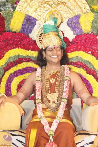 A seated man in a golden crown with a green cloth wrap smiles with eyes closed on a throne-like seat backed by a large arc of red, yellow and purple flower garlands. He wears a rust-brown dress with gold borders, layered gold chain necklaces, a pink and white flower garland, gold armbands and a red bindi. The swept-back long hair, crown, ornate gold necklaces and rudraksha-style styling give probable Swamiji (probable) appearance cues while seated on a flower-decorated throne with a tiger-striped cushion, a gold deity murti or ornament above. Photo of a single in-person figure at a ceremonial darshan setting.