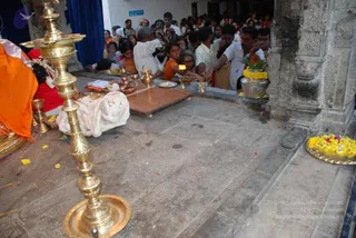 A crowd of devotees presses toward a temple shrine in a stone-pillared hall. In the foreground a tall brass oil lamp stands beside brass vessels and a wooden platform draped with white cloth. On the left, a deity murti wrapped in orange and red cloth with a flower garland is visible. Many people in white, cream and orange clothing lean forward holding brass plates and a lit oil lamp, while a man in a white shirt reaches out. A brass plate of yellow flowers sits on the right beside a kalasha with mango leaves and marigolds. Petals are scattered on the stone floor. The scene reflects a temple puja and darshan during a festival.