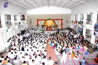 Wide interior photo of a large hall with white walls, a corrugated roof and balconies, filled with a dense crowd seated on the floor, mostly in white clothing, with some in colourful saris on striped mats. At the front, a decorated stage with an orange and gold draped backdrop, a flower-garlanded arch and a central altar with ritual items. Several people in white stand on the stage near the altar, and a figure in purple sits at the front. Fans hang from the walls and ceiling.