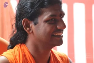 Close-up photograph of a smiling man with long dark hair swept back, wearing a saffron orange shawl draped over his shoulders and a red bindi-style mark on his forehead. Behind him are vertical bands of orange and white. The photo is cropped at the chest and shows only one person, looking off to the side with an open smile.