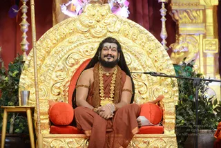 A man seated cross-legged on an ornate gold throne with carved scrollwork and red cushions, smiling with hands folded in his lap. He wears long dark hair, a full beard, a vertical tilak on his forehead, an orange-rust robe, a rudraksha mala and layered gold necklaces, with beaded bracelets on his arms. A microphone stand stands beside the throne, a staff leans at left, and green foliage and a gilded temple backdrop frame the stage. a person cues: long swept hair, rudraksha mala, orange-saffron robes and ornate gold jewellery, seated in a presiding role on the throne.