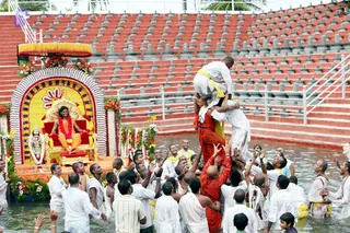 A photograph of a ceremony in a shallow water pool lined with red tiered stone seating. On a decorated golden throne under a canopy with yellow fringe, a figure in orange robes and a red-and-orange garland sits on a red cushion, surrounded by flower garlands and marigold arches. A group of men in white dhotis and yellow sashes, some bare-chested, lift a man in orange robes overhead while others wade in the water with raised hands. Ritual activity is focused around the throne and a deity murti decorated with garlands on the left.
