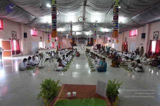 A large hall with white draped fabric ceiling and pink walls hosts a seated meal or ritual gathering. Rows of people sit cross-legged on the polished floor before green banana leaves set with food, while others in white clothes move between the rows. Brightly painted decorated pillars with tassels line the hall, and framed portraits hang on the side walls. Two white cups and a green leaf sit on a red mat in the foreground, flanked by potted green plants. A few people in white kurtas and saris are seated, and a crowd in colourful saris and white clothing fills the middle and far areas near a distant altar.
