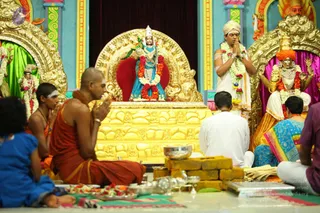 Inside a richly decorated temple shrine, a sannyasi in saffron robes and a bald head sits cross-legged on the floor with hands joined in namaste before a golden throne-like peetha. On the peetha stands a deity murti in blue with red and white garlands. To the right, a man in a white dhoti and shawl stands with palms pressed together, a garland around his neck; a standing figure in white and a seated woman in blue and yellow sari face the shrine. Another seated devotee in a blue top sits at left, a boy in saffron with white tilak sits nearby, and brass vessels, flower offerings, and yellow bricks lie on the floor in front. Golden arches and deity figures frame the shrine, a statue wearing an orange turban and gold ornaments stands at right, and the scene is brightly coloured in turquoise and magenta.