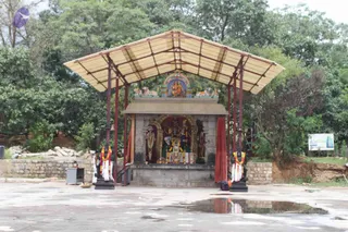 An outdoor shrine under a corrugated pale-yellow canopy supported by red-painted steel posts, set on a wet concrete floor surrounded by green trees. Inside a stone-framed niche stand several painted deity murtis, with a central figure flanked by smaller figures, and a painted panel above showing a seated deity. Two garlanded pillars with marigold garlands stand at the front, a red cloth hangs at the side, and a small offering plate sits on the ground. Stone walls and a signboard appear at the right edge. No people are visible.