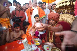A seated man in a saffron-orange turban, orange robe and layered gold and bead necklaces pours water from a copper kalasha over a red ritual table during a puja. Before him stands a small boy with a yellow tilak on his forehead, wearing a white kurta and magenta dhoti, and a second man in a white kurta and orange shawl raises his hand. Behind them, a man in orange with a rudraksha mala films with a video camera, and bare-chested men in orange dhotis watch closely. A golden umbrella and a gold-decorated seat frame the scene, with brass plates of kumkum, turmeric and flower petals on the table.