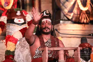A photograph of a bearded man with long dreadlocks seated in front of a decorated altar, raising his right hand in greeting. He wears a leopard-print cloth wrapped around his torso, multiple rudraksha malas in reddish-brown, a gold chain, a silver crescent and small ornament in his hair, white and red forehead markings, and a red cloth at the waist. A microphone stands at right, and behind him are garlands of red roses and white and yellow flowers, a silver-embossed panel, a draped gold textile and a dark deity murti. a person appears to be seated and addressing an audience in a discourse setting.