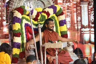 A seated man in saffron-orange robes sits on an ornate silver throne (peetha) decorated with a floral garland of red roses, yellow and white flowers, purple blooms and green leaves. A silver trident (trishul) stands beside him. He wears long dark hair, a beard, a forehead marking and a red wrist thread, and holds a rudraksha mala in his right hand while gesturing with it. A small framed photograph sits on a silver stand in front of him. Bottom of the frame shows the heads of seated devotees, including a child in yellow clothing. The background is a red-gold temple-style backdrop with carved pillars.