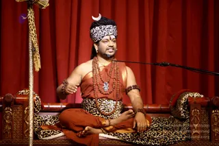 A seated man speaks into a microphone on a wooden throne with carved armrests, upholstered in leopard-print cushions. He wears a leopard-print headband with a crescent ornament, rudraksha malas layered across his chest, a silver pendant, and an orange-red garment with gold border draped over his shoulder, with a matching leopard-print waist cloth. His hands are raised in gesture and he sits in a cross-legged lotus-style posture. Behind him hangs a deep red curtain. Swamiji (probable): the crescent ornament, long swept-back hair, rudraksha malas and ceremonial pendant are visible cues on a person presented in a discourse setting, matching his role as speaker.