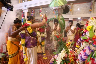 Photograph of a temple ritual inside a hall with decorated pillars and a green-and-gold umbrella overhead. A priest in an orange dhoti and a bare-chested man in a yellow-and-purple cloth hold and lift long garlands of red, yellow, purple and white flowers, with incense smoke drifting nearby. Behind them a man with a long dark braid, a garland of white and pink flowers and folded hands stands in quiet devotion beside another man in white dhoti with hands joined. A woman in a red sari stands at the right among heaps of flowers and leaves on a red floor. A camera rig is visible at the left edge.