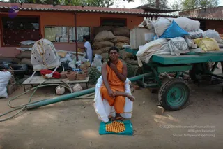 A smiling person in an orange saffron robe sits cross-legged on a yellow and green woven mat in a dusty outdoor yard. A white cloth with a gold border is draped over the lap. Behind them stand a green hand-cart, stacked sacks and cardboard boxes, a small orange-walled building with notice boards, and a person in white walking by. Clay pots and a folded red cushion sit on a wooden table at left. The setting looks like a working ashram compound. No printed name identifies the seated person.