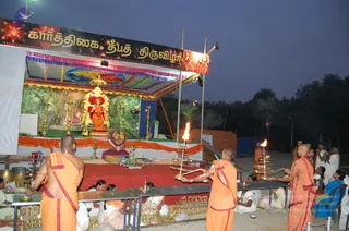 Outdoor evening ritual at a Karthikai Deepam celebration on a raised stage with a banner reading Karthikai Deepam Thiruvizha. Seated on a red-cushioned golden throne at the centre sits a figure in orange robes, a person, with gold necklaces and a turban-like head covering; he is flanked by golden lamps and vases. In front, saffron-robed priests with shaved heads stand holding lit oil lamps and brass vessels, while white-clad women and devotees sit at tables and behind rails. Brass lamps are arranged along the tables, and a tree line and dusk sky sit in the background.
