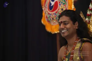 A smiling man with long, swept-back dark hair sits in front of a dark curtain and an ornate orange and red embroidered banner with floral motifs. He wears a red bindi on his forehead, a multi-strand red rudraksha mala, and a green, gold and yellow patterned wrap over bare shoulders. A small white flower garland is visible at the upper right edge. No other people are visible.