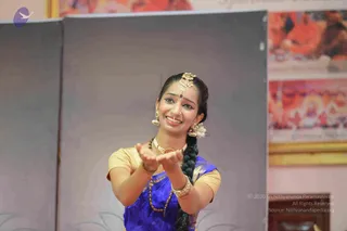 A female dancer in a classical Indian style performs on a stage, smiling with her palms turned upward and held forward. She wears a blue silk sari with gold border, a gold-toned blouse, a gold hair ornament and matching earrings, a flower at her ear, a long braid, and layered beaded necklaces. Behind her, framed panels show saffron-robed figures seated in front of a decorated backdrop, including a deity-style image. The setting appears to be a festival programme or cultural performance.
