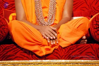 Close crop of a seated figure in saffron-orange robes sitting cross-legged on a red tiger-striped cushioned seat with matching bolsters and a gold-trimmed edge. Clasped hands rest in the lap, and a multi-strand beaded necklace in silver-white hangs on the chest. Only the torso, hands, legs and bare feet are visible; no face or head appears in the frame. The figure's identity cannot be confirmed from the visible cues alone.