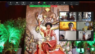 A Zoom screenshot of an online KAILASA session. The main view shows a seated figure in orange-red robes with a long red-and-gold garland, a bead necklace, a black hair crown, and a trident held upright beside him, sitting before a large carved stone relief of a multi-armed deity murti with a cobra-like hood and attendant figures. Green-lit potted plants frame the scene. A grid of six participant tiles at right shows people in home settings: one doing yoga on a mat, another in a blue top and yellow trousers stretching with arms raised, and a woman in a striped top labelled Ma Durga. The Zoom toolbar at top shows Mute, Stop Video, Participants 14, Chat, and a green 'You are screen sharing' banner.