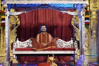 A seated man with long dark hair and a red bindi sits smiling on an ornate silver swing-throne (peetha) with red cushions, wearing rust-orange robes and a saffron shawl over the shoulder, with a microphone stand beside him. Behind him hang red drapes flanked by gilded carved panels and tassels in red, yellow, green and blue. A small golden figurine sits in the foreground. The man's long hair, crown-like swept styling, orange robes and ornate seating are consistent with a probable Swamiji depiction, though his identity is uncertain. The scene appears to be a Sadashivatva session in an ashram hall.