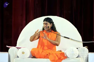 A photograph of a man seated cross-legged on a large white rounded throne against a deep maroon velvet curtain, speaking into a gooseneck microphone. He wears an orange saffron robe draped over his shoulders and a red bindi on his forehead, with long dark hair swept back, and gestures with both hands while talking. Pink papers lie on the throne's side cushions. Appearance-based candidate: a person, seated and addressing an audience on a stage.