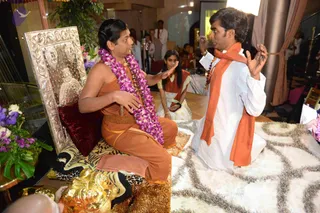 Photograph of a seated figure on an ornate silver-and-gold throne draped with a black-and-gold tiger-stripe cloth and a maroon cushion. The seated person wears an orange-saffron dress and a long purple orchid garland, with a rudraksha mala visible at the chest, and gestures with raised open hands toward a kneeling man. The kneeling man in a white kurta and orange shawl with a rudraksha mala gestures expressively with both hands, facing the seated figure on the white shag rug. Behind them a woman in a white and orange sari sits, and other people in white and orange clothing stand near a stage with lamps and flower arrangements. A gilded lion or tiger figure is visible at the lower edge. The seated figure has long swept-back hair and orange ceremonial clothing, which is consistent with a probable Swamiji (probable) presence in the scene.