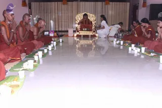 A hall with a polished white floor where a seated figure in a red-orange robe and rudraksha mala sits on an ornate gold throne at the far end. Lines of devotees in red-orange and saffron clothing sit with hands folded in anjali on both sides of the floor. Banana-leaf plates with food, cups and flowers are laid out along the lines. A woman in a white sari kneels near the throne and attends to metal vessels, while another woman in white works at the right. A framed photograph sits near the left side. Setting suggests a ritual meal and puja gathering.