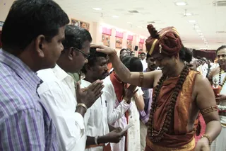 Indoor photo of a hall with pale walls and ceiling lights, where a woman in an orange-brown sannyasi wrap, rudraksha mala and a rust-coloured turban touches the forehead of a devotee in a white shirt with a raised hand, with a smiling expression. Around her, men and women in white and light clothing stand with palms pressed together in namaste, some holding folded paper. Framed pictures hang on the wall behind and a person in white stands on the right, with a rudraksha mala. A brass vessel is visible at the right edge. The scene suggests blessing or darshan. The woman is a sannyasi-attired figure, not a confirmed a person, so identity is uncertain.