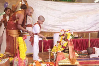 A photograph of a ritual scene under a white cloth canopy. A shaved-headed priest in an orange-brown dhoti and shawl, with a rudraksha mala, leans forward and guides a young shaved-headed boy in white clothes and an orange sash as the boy reaches toward a floral-garlanded gold-edged platform. Yellow and orange marigold garlands and a pink cloth drape the platform, with brass oil lamps nearby and a flower-decked tray in the foreground. At right, a man in white lies prostrate on red and patterned mats. Other white-clad attendants stand behind. The setting suggests a homa or samaya deeksha ceremony.