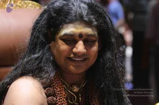 Close-up photo of a smiling person with long, dark, wavy hair and a yellow-and-brown forehead marking with a dark bindi, seated in front of a brown leather chair with gold ornamental trim. The person wears a dense rudraksha mala of reddish-brown beads and a gold pendant necklace, and is bare-shouldered. Background is blurred with other people in muted clothing.