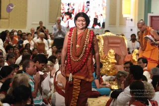 A standing figure in the centre of a seated audience in a bright hall walks forward smiling, with long dark swept-back hair, a red bindi and a shawl of rust-red and saffron cloth over a rust-red garment. Around the neck hang layered garlands of red roses and white flowers, a rudraksha mala and a gold pendant necklace. Behind and to the right, a robed priest in orange and saffron stands holding an open book, beside a gold and cushioned throne. Devotees seated on the floor press their palms together in anjali, and a large screen above shows more of the crowd.