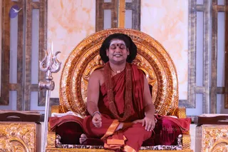 A seated figure in red-maroon robes sits cross-legged on an ornate gold throne with a large carved backrest against a marble-patterned wall. He wears a white tilak with a red dot on the forehead, a rudraksha mala, a gold necklace, and an orange-trimmed dhoti, with a bracelet on his wrist and hands in a mudra. A silver trident stands to his left beside a carved gold side table, and a maroon cushion rests behind him. Probable Swamiji cues include long swept-back hair, rudraksha malas and ceremonial gold necklace while seated on the throne.