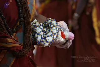 Close photograph of a person in a dark red garment with a gold-edged border holding a silver offering tray. The tray carries white jasmine-style flower garlands strung with blue beads and gold accents, plus a small orange flower and a pink lotus-like bloom. A thick rudraksha mala with dark seeds hangs across the torso. Blurred figures in matching red robes stand in the background. A small purple bird logo appears at the top left. No face is shown, so no a person identification is made.