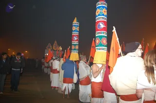 Night procession on a misty road at a Kumbh Mela-style event. Robed participants in white dhotis and orange-red waistcloths walk barefoot, carrying tall cylindrical painted banners decorated with floral patterns, geometric bands and white dove emblems, alongside long orange flags. A man in the foreground at right wears a white shawl and a white head covering and holds a staff; a figure at the front with a bare upper body carries a banner. Several spectators in dark jackets stand at the left. Orange-yellow street lights glow through the haze. Whether a person in white is a person cannot be confirmed from appearance alone, so the identity is left unsure.