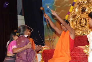 A seated man in an orange-saffron robe over a red top sits on a red patterned cushioned seat before an ornate gold throne with carved deity relief and yellow-and-purple flower garlands. He raises a trident in one hand and a wooden staff in the other. Before him, a woman in a purple patterned sari with a gold-bordered shawl and a woman in a pink blouse stand with hands folded in respect. A man in an orange robe stands behind them. Dry twigs lie in front of the throne. Appearance cues including the saffron robe and throne are consistent with a person, seated in a blessing gesture with a trident.