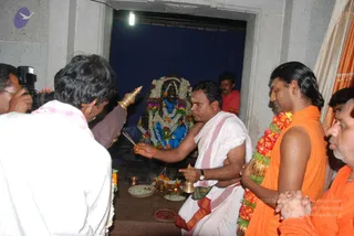 A photograph inside a small shrine with a dark blue cloth backdrop and a deity murti decorated with garlands of white and orange flowers. A bare-chested priest in white dhoti and shawl with red border holds a bell and incense, performing puja. Beside him, a man in saffron-orange robes with a long garland of orange and red flowers stands with hair swept back and gold bangles on the wrist. A cameraman in white shirt and a man in orange clothing stand at the edges. Brass lamps, offering plates and flower petals sit on the altar.