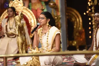 A woman in a cream and gold-bordered kerala-style sari sits on the floor speaking into a handheld microphone during a satsang. She wears a gold necklace with beads, long gold earrings, a red bindi and glasses. Beside her, a man in white dhoti and shawl with a rudraksha mala sits attentively. Behind them a golden deity murti stands on the left, with a golden kuthuvilakku oil lamp in front, and a seated woman in white and gold sits in the background near an ornate golden arch and lit lamps. The setting is a stage with gold ornamentation and a decorated carpet.