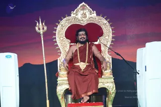 A man with long dark hair and a full beard sits on an ornate gold throne with a red velvet back and seat, on a stage with a painted sunset-over-mountain backdrop. He wears a rust-red robe over the shoulders, a long gold-beaded mala necklace, a gold waist belt, and gold bracelets, with a red tilak on his forehead. A golden trident stands to his left, and a microphone stands to his right beside white air-conditioning units.